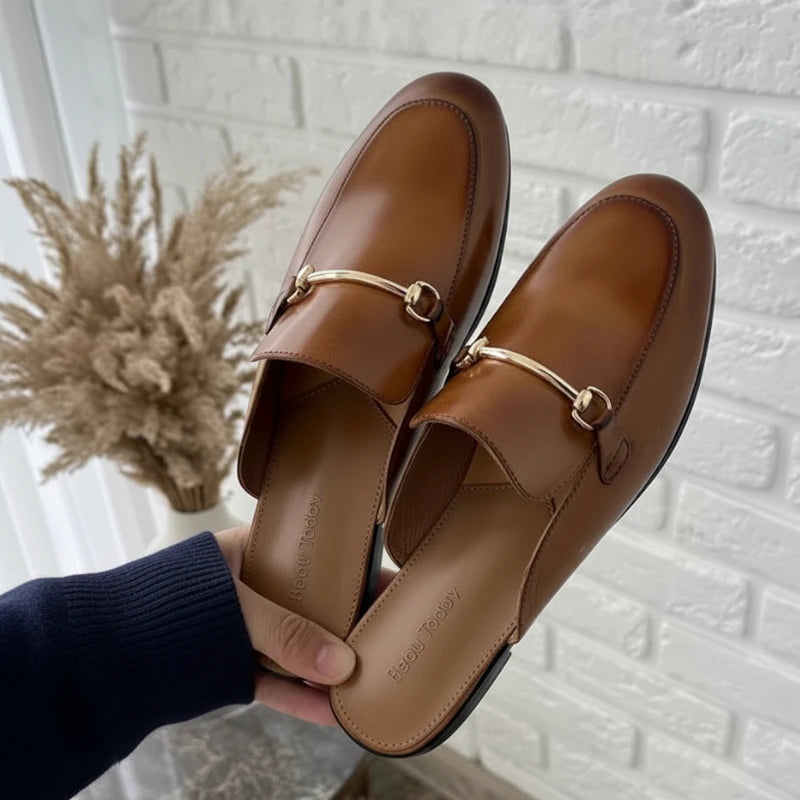 Paire de Mules Femme Chic en Cuir Marron tenue en main, montrant la brillance du cuir véritable et l’ornement métallique sur le dessus.