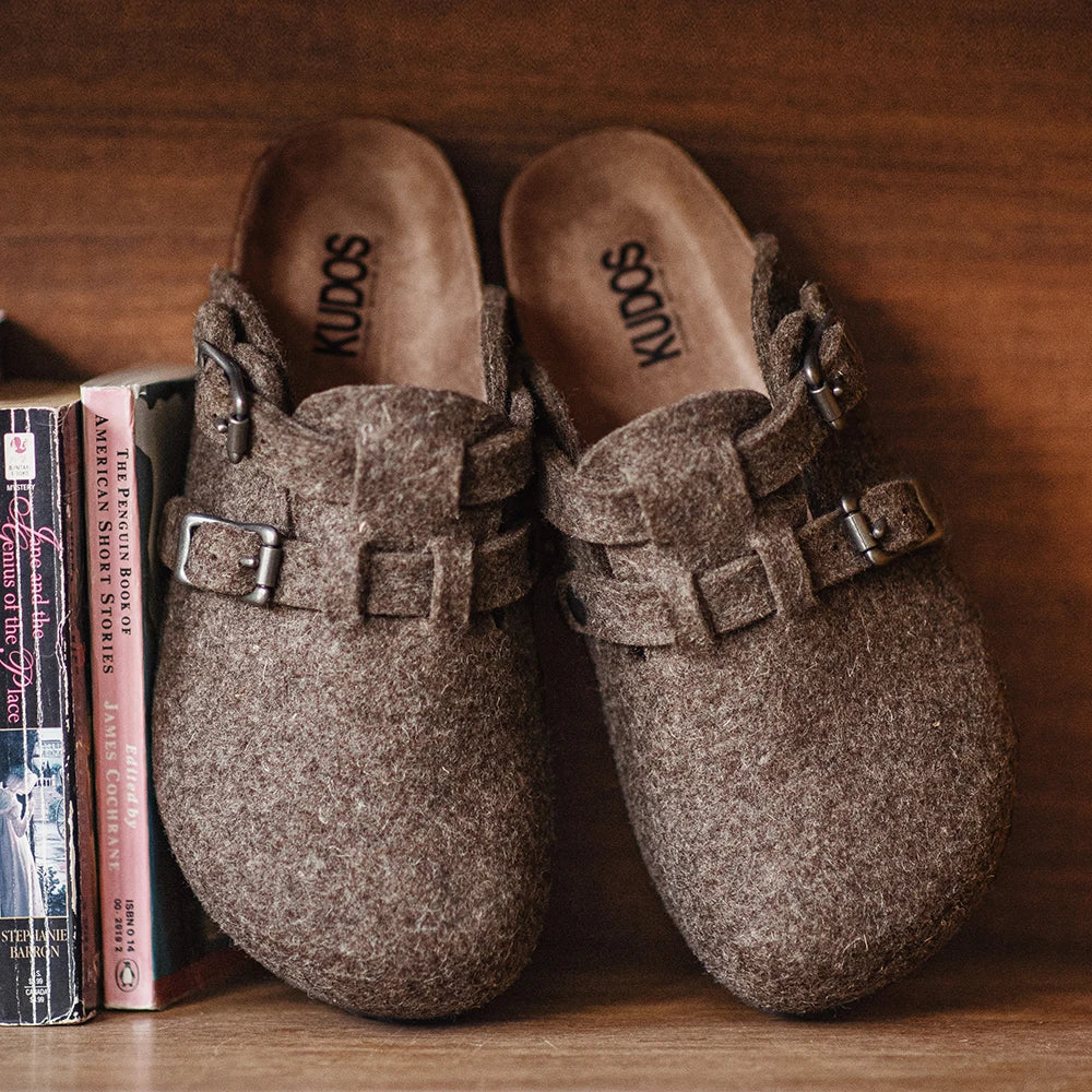 Mules marron vintage en laine feutrée double boucles métallique posées sur étagère bois foncé à côté livres semelle liège visible